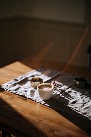 A white cup filled with coffee sits on a wooden table. The table is partially covered with a white cloth, and small decorative items are scattered around. Soft lighting creates gentle shadows, adding a warm and inviting atmosphere.
