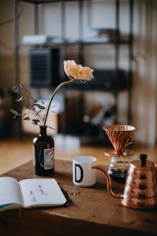 A cozy kitchen scene with a steaming cup of coffee beside an open notebook and a vase of fresh flowers.