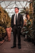 A man in a dark suit stands confidently in a greenhouse surrounded by lush green plants. The pathway is lined with potted foliage, and a structure made of glass and metal frames arches overhead.