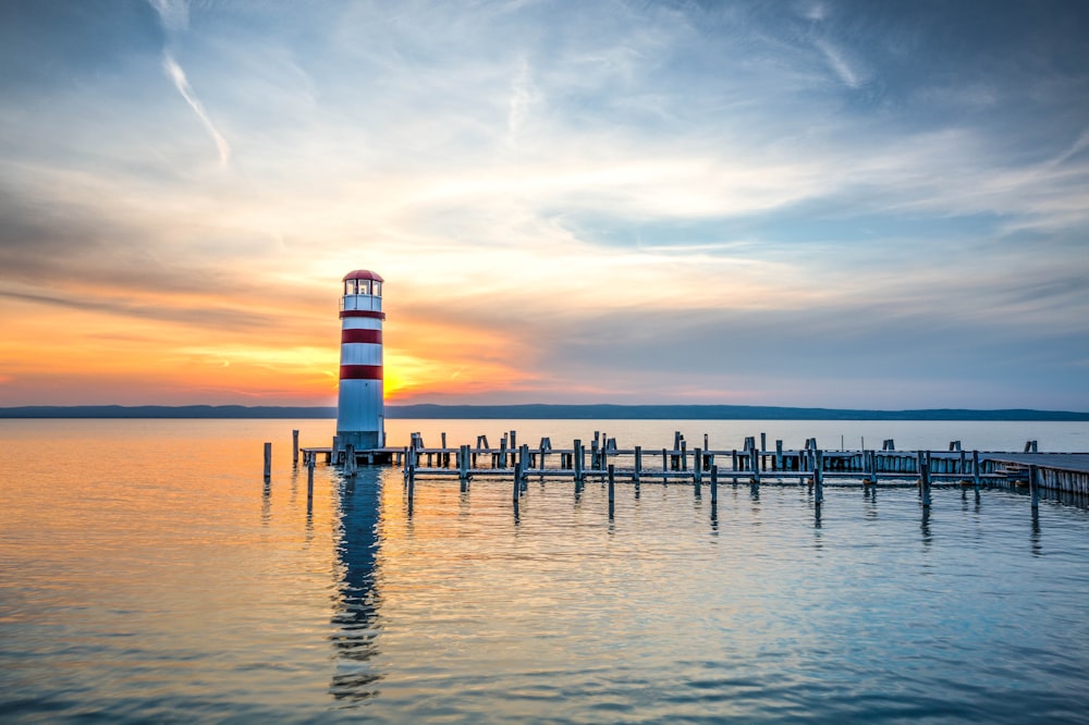 photo of white and red lighthouse beside wooden beach dock under clear blue sky