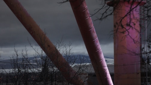 Industrial pipes or structures with a pinkish hue against an overcast sky. Bare tree branches are visible in the foreground and there are silhouettes of mountains in the background.