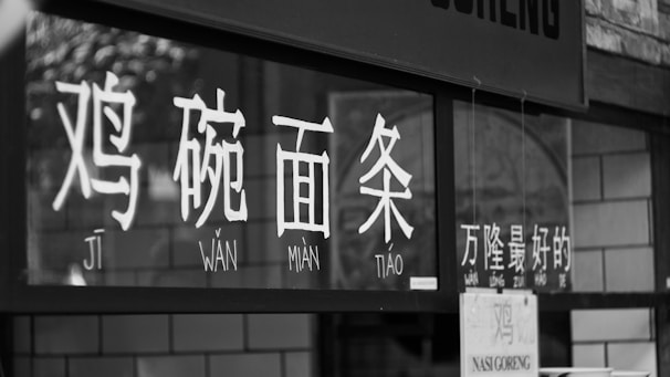 A black and white close-up of a store or restaurant sign displaying Chinese characters with accompanying pinyin. The characters seem to be printed on a window or glass surface.