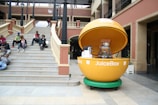 A large, yellow, spherical JuiceBox with an open top is prominently displayed. It resembles a giant orange and contains juicing equipment. Adjacent to a wide stone staircase, several people are sitting and relaxing. The setting appears to be a modern outdoor plaza with a two-story building featuring brick and beige elements.
