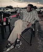 An elderly man is sitting comfortably in a foldable camping chair on the side of the road. He is wearing a patterned shirt, beige trousers, and sneakers with a unique design. Around him are several parked cars, and the sky is overcast, suggesting a relaxed atmosphere.