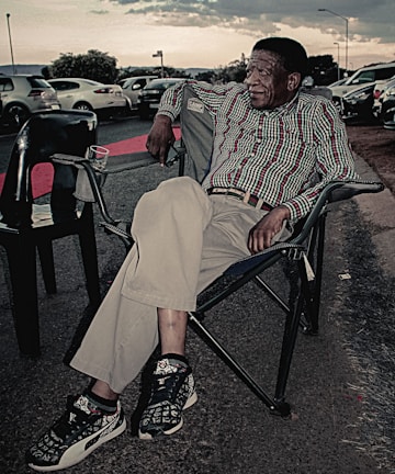 An elderly man is sitting comfortably in a foldable camping chair on the side of the road. He is wearing a patterned shirt, beige trousers, and sneakers with a unique design. Around him are several parked cars, and the sky is overcast, suggesting a relaxed atmosphere.