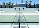There is a tennis court with pale green surfaces and white boundary lines in a sunny outdoor environment. Palm trees and a white building are visible in the background against a backdrop of mountains under a clear blue sky. The scene is symmetrical with empty seating areas on either side.
