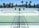 There is a tennis court with pale green surfaces and white boundary lines in a sunny outdoor environment. Palm trees and a white building are visible in the background against a backdrop of mountains under a clear blue sky. The scene is symmetrical with empty seating areas on either side.