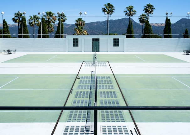 There is a tennis court with pale green surfaces and white boundary lines in a sunny outdoor environment. Palm trees and a white building are visible in the background against a backdrop of mountains under a clear blue sky. The scene is symmetrical with empty seating areas on either side.