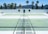 There is a tennis court with pale green surfaces and white boundary lines in a sunny outdoor environment. Palm trees and a white building are visible in the background against a backdrop of mountains under a clear blue sky. The scene is symmetrical with empty seating areas on either side.