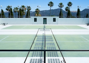 There is a tennis court with pale green surfaces and white boundary lines in a sunny outdoor environment. Palm trees and a white building are visible in the background against a backdrop of mountains under a clear blue sky. The scene is symmetrical with empty seating areas on either side.