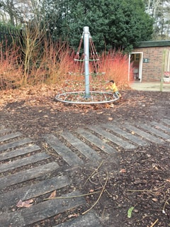 A playground with a rope climbing structure surrounded by fallen leaves and red autumnal shrubs. A child in a yellow jacket is visible beside the climbing frame. The area is bordered by tall evergreen shrubs and a brick building with an open door.