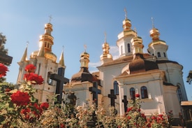A beautiful orthodox church with gold-domed towers shines brightly against a clear blue sky. Vibrant red roses and dark crosses line the foreground, enhancing the serene atmosphere.