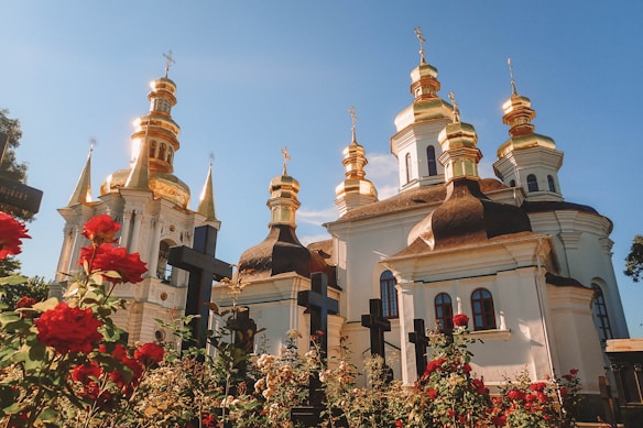 A beautiful orthodox church with gold-domed towers shines brightly against a clear blue sky. Vibrant red roses and dark crosses line the foreground, enhancing the serene atmosphere.