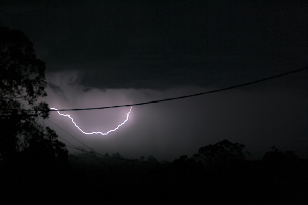A powerful lightning flash illuminating a rural landscape with silhouetted trees.