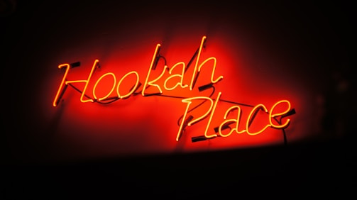 A neon sign with bright orange and red colors displays the words 'Hookah Place' against a dark background. The glowing letters create a vibrant and inviting atmosphere.