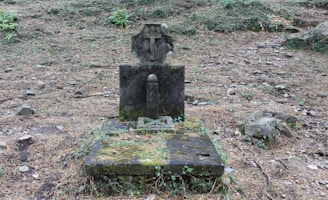 An old, weathered gravestone with a cross on top stands on a moss-covered stone base. It is surrounded by a sparse, rocky, and overgrown area with patches of grass and fallen leaves.
