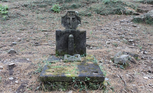 An old, weathered gravestone with a cross on top stands on a moss-covered stone base. It is surrounded by a sparse, rocky, and overgrown area with patches of grass and fallen leaves.