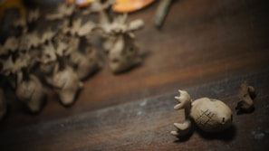 Small clay sculptures resembling reindeer with antlers are scattered across a wooden surface. A single reindeer figurine is prominently in focus, displaying carved patterns on its body.