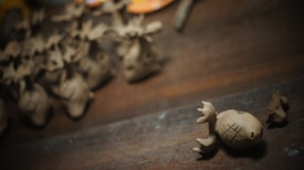 Small clay sculptures resembling reindeer with antlers are scattered across a wooden surface. A single reindeer figurine is prominently in focus, displaying carved patterns on its body.
