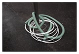 A green hose is coiled around a gray pole with several white loops intersecting it. The background consists of a rough, dark textured surface, possibly asphalt.