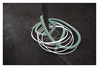 A green hose is coiled around a gray pole with several white loops intersecting it. The background consists of a rough, dark textured surface, possibly asphalt.