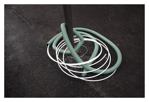 A green hose is coiled around a gray pole with several white loops intersecting it. The background consists of a rough, dark textured surface, possibly asphalt.