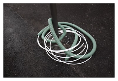 A green hose is coiled around a gray pole with several white loops intersecting it. The background consists of a rough, dark textured surface, possibly asphalt.