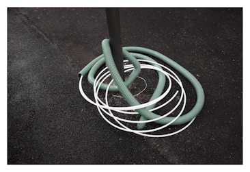 A green hose is coiled around a gray pole with several white loops intersecting it. The background consists of a rough, dark textured surface, possibly asphalt.