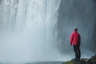 A person in a red jacket stands on a rocky outcrop, gazing at a massive waterfall with mist billowing all around. The scene captures a feeling of awe and natural grandeur.