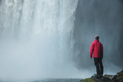 A person in a red jacket stands on a rocky outcrop, gazing at a massive waterfall with mist billowing all around. The scene captures a feeling of awe and natural grandeur.