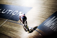 A cyclist wearing a white and black uniform is riding a track bike on an indoor velodrome. The wooden track is visible, with shadow and light creating a dynamic pattern. Portions of the track have large lettering printed on them.