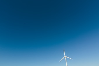 A solitary wind turbine stands against a vast expanse of clear blue sky, its white blades gently angled. The simplicity and openness of the landscape create a serene and modern atmosphere.