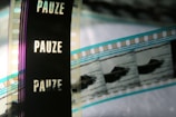 A close-up of a filmstrip with several frames, partially featuring the word 'PAUZE' vertically against a dark background. The image also shows segments of the film containing images, possibly of a boat or similar object, with the filmstrip showing shades of blue, black, and transparent sections.