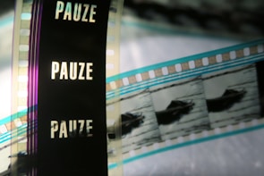 A close-up of a filmstrip with several frames, partially featuring the word 'PAUZE' vertically against a dark background. The image also shows segments of the film containing images, possibly of a boat or similar object, with the filmstrip showing shades of blue, black, and transparent sections.