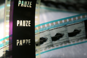 A close-up of a filmstrip with several frames, partially featuring the word 'PAUZE' vertically against a dark background. The image also shows segments of the film containing images, possibly of a boat or similar object, with the filmstrip showing shades of blue, black, and transparent sections.