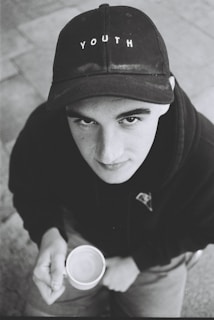 A young person gazes upward while holding a cup. They are wearing a black cap with the word 'YOUTH' on it and a dark hoodie. The image is in black and white, contributing to a nostalgic or contemplative atmosphere.