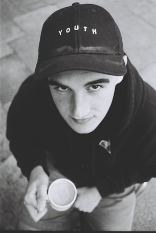 A young person gazes upward while holding a cup. They are wearing a black cap with the word 'YOUTH' on it and a dark hoodie. The image is in black and white, contributing to a nostalgic or contemplative atmosphere.