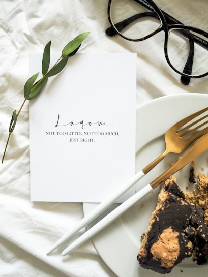 A minimalistic setup features a white card with the word 'Lagom' and a phrase below it, placed on a white fabric background. Next to the card, a sprig of greenery is visible. A pair of black-rimmed glasses is positioned beside the card. Adjacent to the card, a white plate holds a piece of cake, accompanied by a gold fork and knife.