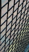 A close-up view of a black metal wire mesh fence with a blurred background. The fence bars are interwoven in a diamond pattern, and the background appears to be a paved area with a subtle mix of blue and earthy colors.