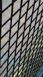 A close-up view of a black metal wire mesh fence with a blurred background. The fence bars are interwoven in a diamond pattern, and the background appears to be a paved area with a subtle mix of blue and earthy colors.