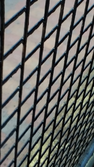 A close-up view of a black metal wire mesh fence with a blurred background. The fence bars are interwoven in a diamond pattern, and the background appears to be a paved area with a subtle mix of blue and earthy colors.