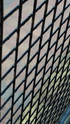 A close-up view of a black metal wire mesh fence with a blurred background. The fence bars are interwoven in a diamond pattern, and the background appears to be a paved area with a subtle mix of blue and earthy colors.