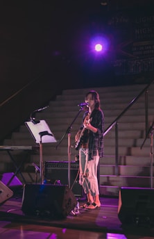 A musician is performing on stage with a microphone and an electric guitar. The setting is dimly lit with a spotlight in the background, creating a dramatic ambiance. There are musical instruments and equipment around, including a keyboard and speakers.