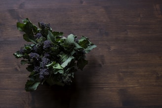 A bunch of leafy greens with dark purple florets is placed on a wooden surface, with the focus on the textured leaves and florets.