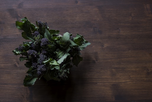 A bunch of leafy greens with dark purple florets is placed on a wooden surface, with the focus on the textured leaves and florets.