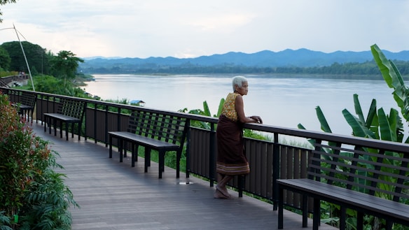An elderly person with short white hair stands on a wooden boardwalk overlooking a wide river. The person leans on a railing, appearing contemplative. The boardwalk is lined with benches and lush greenery, including banana plants. In the distance, there are rolling hills and a cloudy sky, creating a serene backdrop.