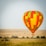 A colorful hot air balloon floats over a grassy savanna landscape. The balloon is prominently red, orange, yellow, and green, and features the words 'Balloon Adventure' on its surface. Below, a safari vehicle is parked in the tall grass, and there are scattered trees in the background under a clear sky.