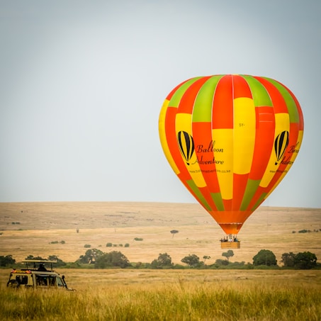A colorful hot air balloon floats over a grassy savanna landscape. The balloon is prominently red, orange, yellow, and green, and features the words 'Balloon Adventure' on its surface. Below, a safari vehicle is parked in the tall grass, and there are scattered trees in the background under a clear sky.