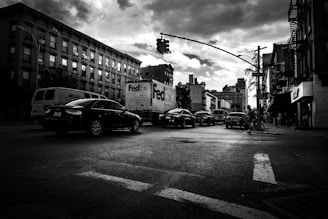 A bustling urban street scene with vehicles including a FedEx truck and a bicycle. Buildings line both sides of the street, with a traffic light overhead. The sky appears cloudy, casting dramatic shadows on the street.