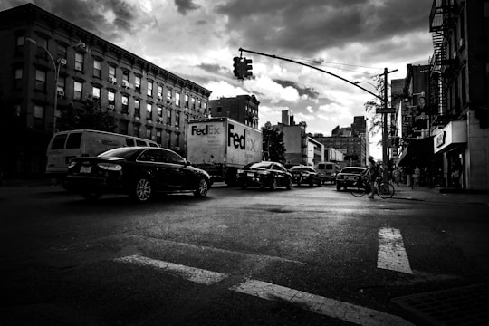 A bustling urban street scene with vehicles including a FedEx truck and a bicycle. Buildings line both sides of the street, with a traffic light overhead. The sky appears cloudy, casting dramatic shadows on the street.